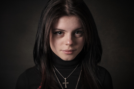Young woman with long dark hair wearing a black turtleneck and silver cross necklaces against a dark background.