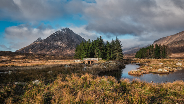 Mountain peak rises above a moorland valley with a stream, conifer forest, and golden grasses under a partly cloudy sky.
