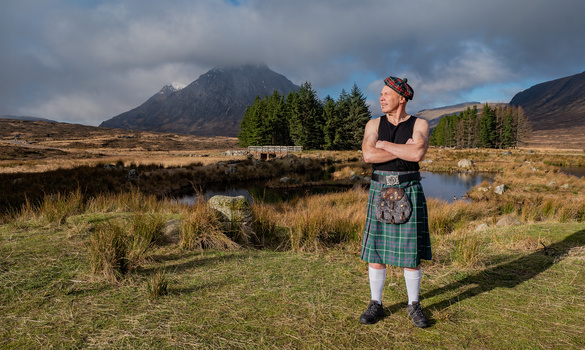 Man in traditional Scottish kilt standing in moorland valley with mountain backdrop.