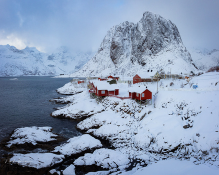 Red wooden houses on snowy shore beneath steep snow-covered mountain peak.