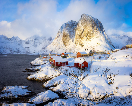 Snow-covered coastal village with red cabins nestled beneath a dramatic rocky peak in a winter fjord landscape.
