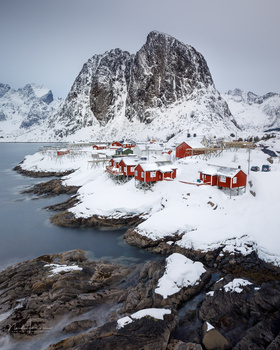 Snow-covered coastal village nestled beneath a dramatic jagged mountain peak, with red cabins and boats on a frozen fjord.