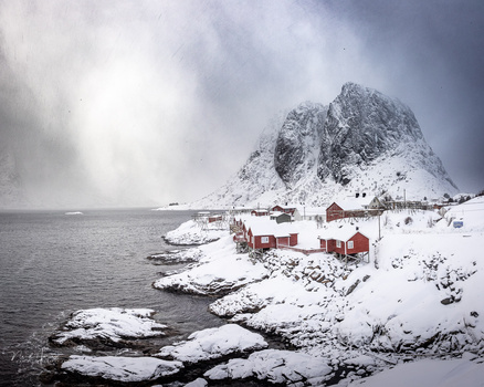 Snow-covered coastal village with red buildings nestled below a dramatic rocky peak rising from the sea.