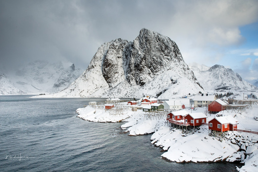 Snow-covered fishing village nestled beneath a dramatic peaked mountain on a fjord coastline.