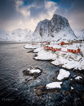 Snow-covered mountain peak rising above a frozen fjord with red cabins and fishing boats nestled along the icy shoreline.