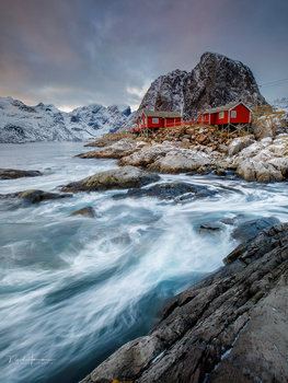 Red cabin perched on rocky coastline with turbulent sea and snow-capped mountains beyond.