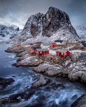 Red wooden cabins nestled on rocky shores beneath a dramatic snow-capped mountain peak.
