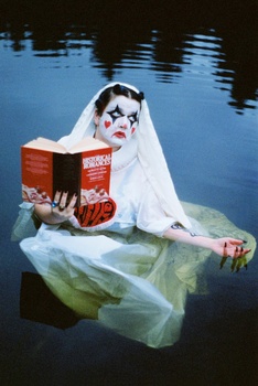 Person in white veil and theatrical makeup holding red book while floating in transparent plastic over still water.
