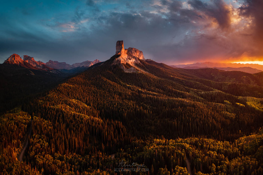 Mountain peak illuminated by golden sunset light, surrounded by forested slopes under dramatic cloud-streaked sky.