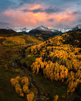 Golden larch trees blanket a valley floor beneath snow-capped mountains under a dramatic sunset sky.