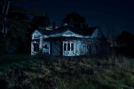 Abandoned residential house photographed at night with blue moonlight casting stark shadows across overgrown grass.
