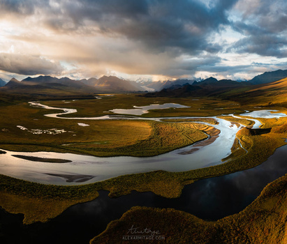 Aerial view of a meandering river through tundra with distant mountains under dramatic cloudy sky.