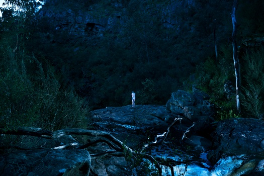 Solitary figure standing on moss-covered rocks in a dark cave opening at dusk.
