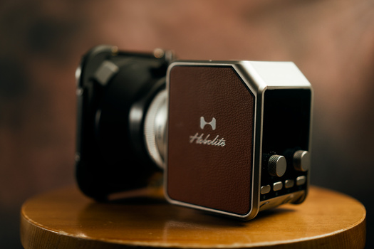 Brown and black compact camera accessory with chrome trim displayed on wooden surface, with DSLR camera blurred in background.