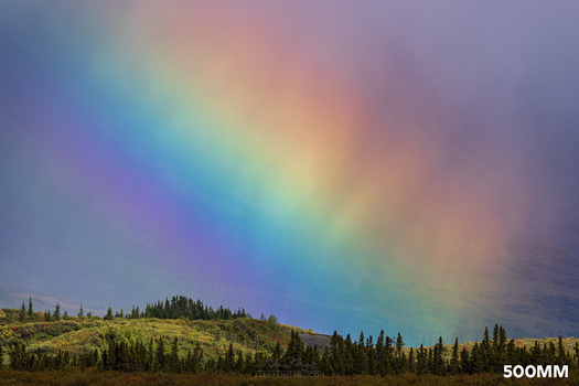 Vivid rainbow arc stretching across a stormy sky above a forested mountain landscape.