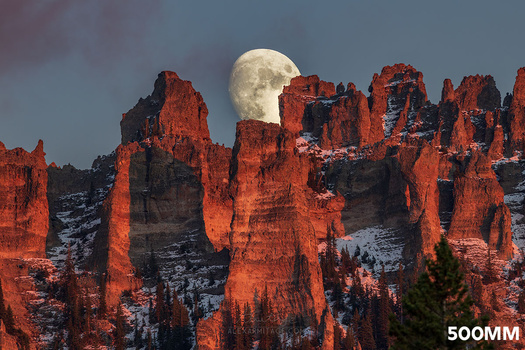 Full moon rising behind towering red rock formations dusted with snow.