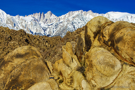 Golden boulders in foreground with snow-capped mountain range under blue sky in background.