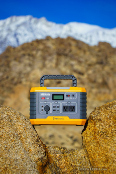 Portable power station with yellow and gray casing positioned on desert rocks with snow-capped mountains in background.