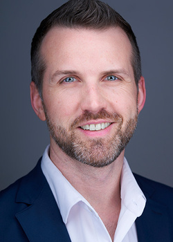 Professional headshot of a man with beard wearing dark blazer and white shirt against neutral gray background.