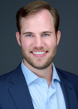 Professional headshot of a man with light brown hair and beard wearing a navy blazer and light blue shirt against a neutral gray background.