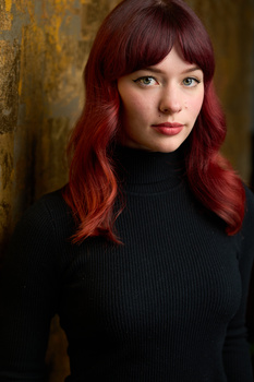 Woman with burgundy hair and bangs wearing a black turtleneck against a textured gold and brown background.
