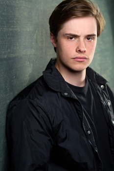 Young man in dark jacket against textured green-gray backdrop, direct gaze and neutral expression.