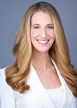Woman with long blonde wavy hair smiling at camera wearing white blazer.