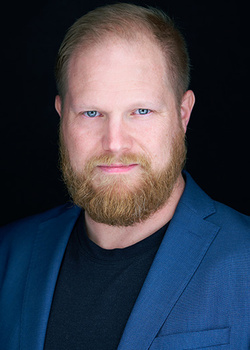 Professional headshot of a man with reddish-brown beard wearing a blue blazer over a black shirt against a dark background.