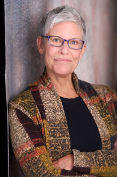 Portrait of a woman with gray hair and glasses wearing a patterned jacket.