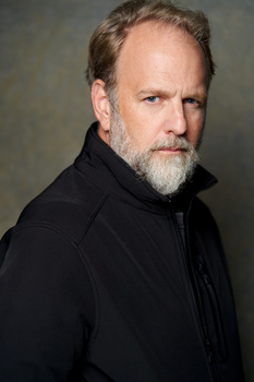 Man with beard wearing black jacket against neutral gray background.