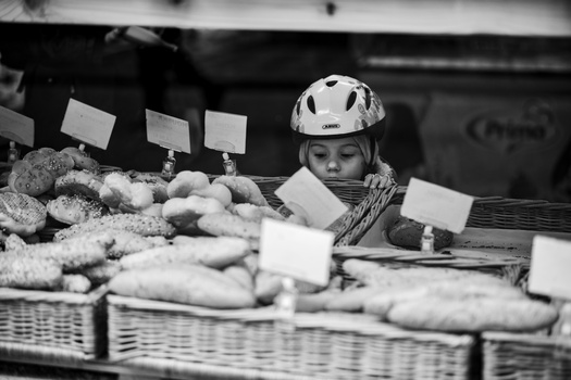 Child wearing a bicycle helmet sits behind a market display of fresh produce with price cards.