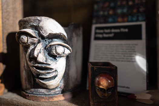 Sculptural head with exaggerated facial features and wearing glasses, displayed in a gallery setting with descriptive placard and additional artwork visible in background.