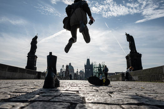 Silhouetted figure airborne above a stone bridge with historic city buildings reflected in calm water below.