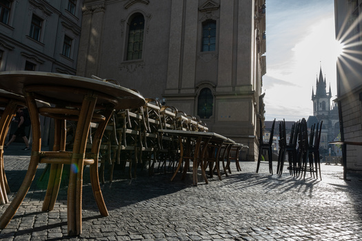 Empty outdoor café seating with stacked chairs in bright sunlight, historic architecture and church spire visible in background.