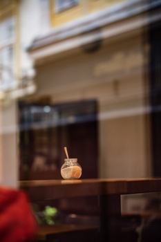 Iced beverage in a glass with straw positioned on a dark wooden table, with blurred storefront and red fabric in foreground.