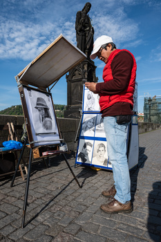 Street portrait artist in red jacket displaying framed sketches at outdoor market beneath statue.