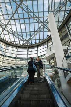 Two people riding an escalator upward inside a modern glass-roofed atrium with geometric steel framework.