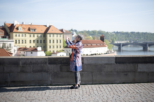 Person in patterned dress holding up a smartphone to photograph a riverside cityscape with historic buildings and a bridge.