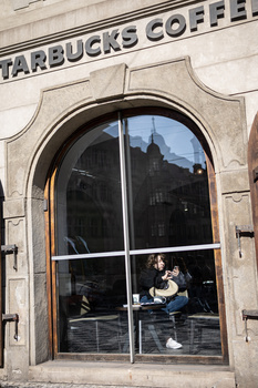 Person sitting inside a Starbucks storefront window, viewed through glass panes with architectural framing.