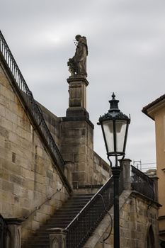 Stone staircase with ornate statue on pedestal and vintage street lamp against overcast sky.