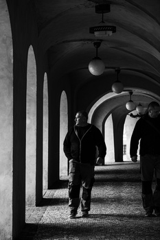 Solitary figure standing in a columned corridor with dramatic architectural shadows and overhead lighting.