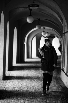 Man standing in a long arched corridor with repeating architectural elements and dramatic shadows.