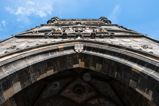 Ornate stone archway with decorative moldings and a shield emblem at its apex, photographed from below against a blue sky.