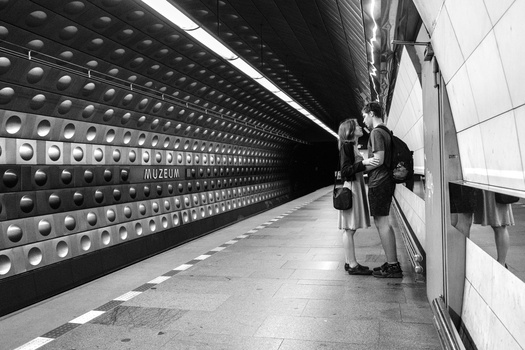 Two people standing on a subway platform with distinctive circular perforated wall paneling behind them.