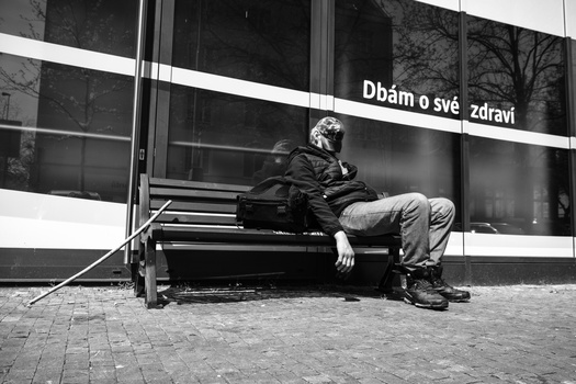 Man sitting on a bench against a modern building with Czech text on its facade, shot in black and white.