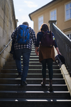 Two people walking up outdoor stairs toward a yellow building on a bright day.