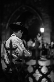 Man in fedora and light shirt standing in shadowed archway at night, blurred figures in background.