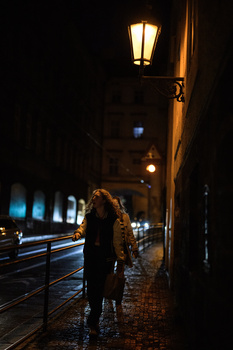 Person standing on wet cobblestone street at night under a glowing street lamp with blurred car light trails in background.