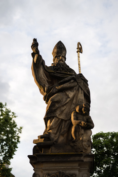 Bronze statue of a robed religious figure holding a staff and raising one hand, photographed against a cloudy sky.