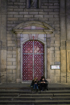 Two people sitting on stone steps in front of a large ornate wooden door with decorative ironwork.